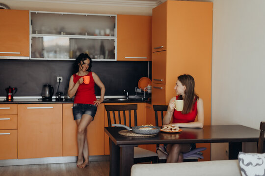 Two women spend time in modern kitchen with one holding warm drink near counter while another sits at table creating lifestyle scene about daily rituals social connection home comfort and slow living.