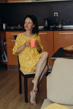 Woman in yellow dress sits barefoot with orange mug in modern kitchen creating quiet lifestyle scene around digital detox reflection slow living mindful mornings emotional balance and home comfort.