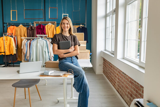 A young woman stands confidently in her small clothing business, holding a tablet. The store features colorful garments displayed on racks and boxes stacked nearby. Natural light floods the space, cre