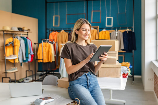 A young woman smiles while using a tablet to manage her small clothing business. She sits on a table surrounded by cardboard boxes and colorful clothing displays in a bright, modern boutique.