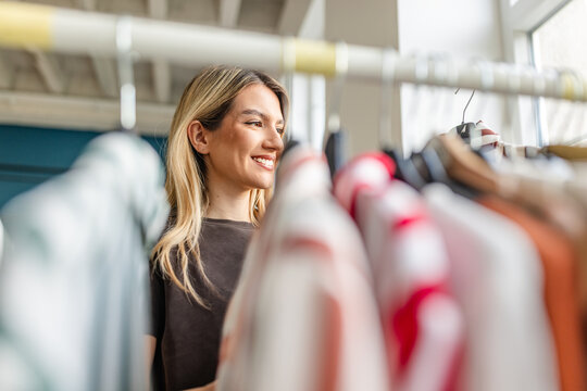 A young woman browses through a rack of colorful clothing in a boutique. She smiles as she examines the garments, enjoying the shopping experience in a bright, modern setting.