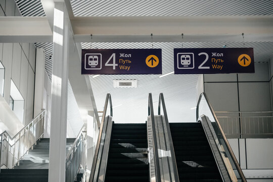 Directional signs above escalators showing railway platform numbers at a train station in Aktobe, Kazakhstan.

