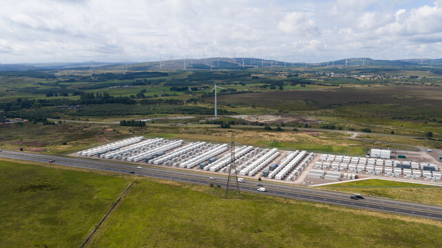 Aerial view of a large battery energy storage system with rows of white containers next to a highway in a rural landscape with wind turbines in Coalburn, Scotland, United Kingdom.
