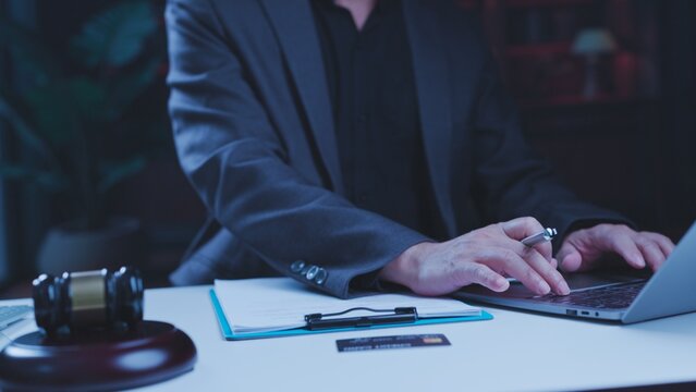 Corporate lawyer typing laptop computer beside wooden gavel clipboard credit card working late night dark office reviewing legal financial contract documentation.