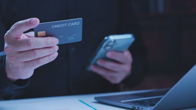 Hand holding credit card next smartphone near laptop desk under low light, ready online payment transaction, digital banking authentication, e-commerce checkout, financial authorization.