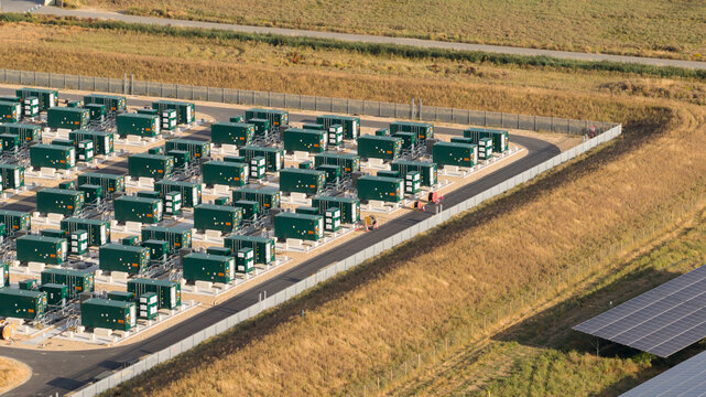 Aerial view of a battery energy storage system facility with rows of green containerized battery units, gravel ground, and perimeter fencing in London, England, United Kingdom.