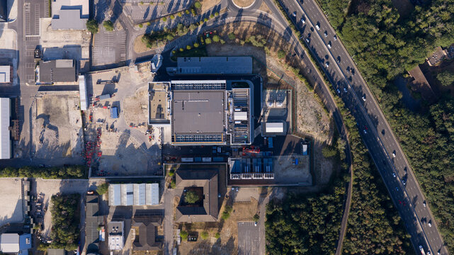 Aerial view of a large industrial facility with a flat roof, surrounding parking areas, and construction zones, situated adjacent to a multi-lane highway in Longcross, England, United Kingdom.