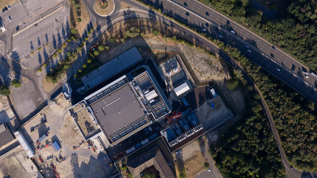 Aerial view of a large industrial facility with a flat roof, parking areas, and adjacent highway in Longcross, England, United Kingdom.