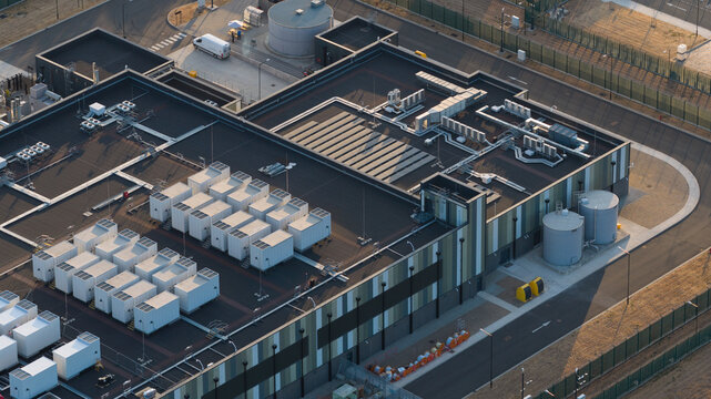 Aerial view of a modern data center facility featuring rows of white cooling units on the roof, industrial storage tanks, and a secure perimeter in Bracknell, England, United Kingdom.