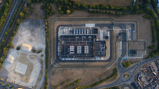 Aerial view of a modern data center facility featuring rooftop cooling units and secure perimeter fencing, situated near roads and residential areas in Bracknell, England, United Kingdom.