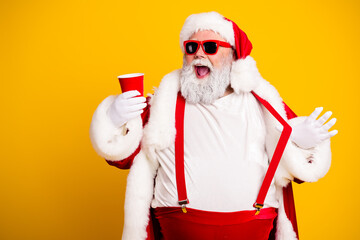 Obraz premium Cheerful Santa Claus in sunglasses holding a red party cup celebrating Christmas and holiday fun in a festive red and white outfit against a bright yellow background