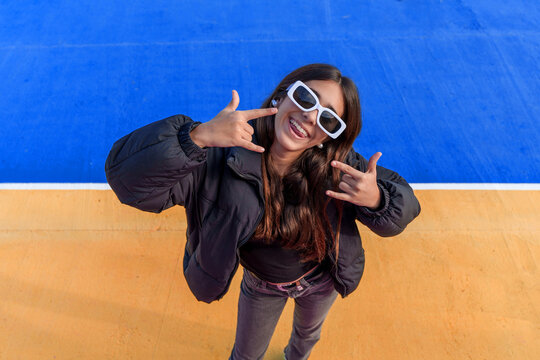 Young smiling girl wearing sunglasses making rock and roll hand gesture against a colorful blue and orange background