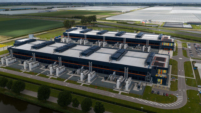 Aerial view of a large modern data center facility with industrial cooling units on the roof, surrounded by green fields and greenhouses in Middenmeer, North Holland, Netherlands.