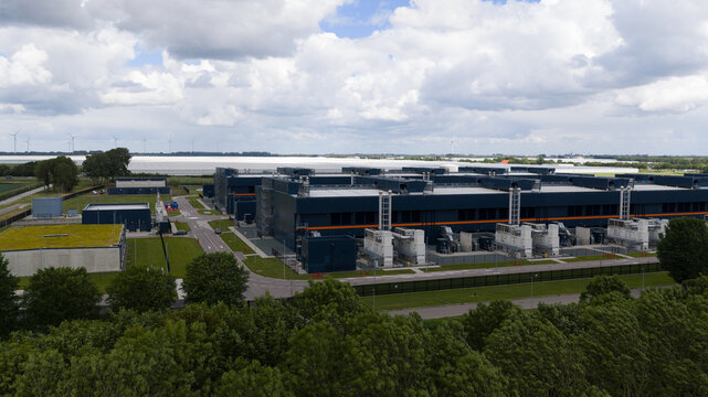 Aerial view of a large modern data center with industrial buildings, cooling units, and a green roof, surrounded by trees and wind turbines in Middenmeer, North Holland, Netherlands.