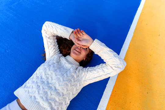 Young woman smiling while lying on a vibrant blue and yellow court, shielding her eyes with hands. Relaxed outdoor portrait