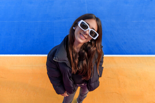 Young woman wearing white sunglasses smiling, looking up at camera against a colorful blue and orange background