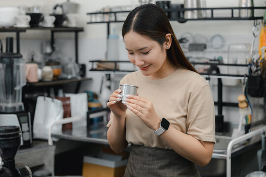 Asian barista smelling coffee aroma from metal cup in modern cafe, highlighting quality control, tasting, espresso preparation, small business, hospitality service, workplace lifestyle.