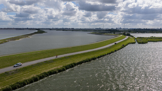 Aerial view of a dike road separating water bodies with a car and a cyclist on the road, lush green grass, and a distant city skyline in Uitdam, Netherlands.