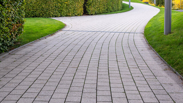 Wide perspective of a paved stone walkway curving through manicured green lawns and trimmed hedges. Neat pedestrian path design suitable for urban parkland, greening and landscaping