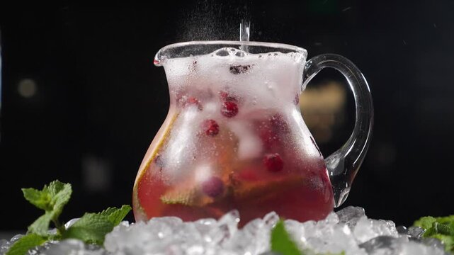 Water pouring from above into glass pitcher with berry lemonade, ice, mint and red berries. Splashes and bubbles, dark background, refreshing summer drink. Professional photography. Slow motion hd