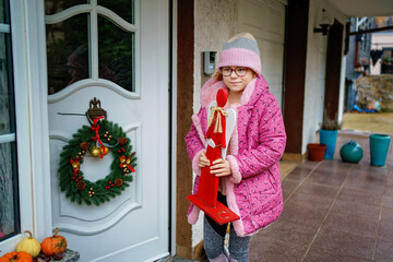 Obraz premium Little girl standing at the front door holding Christmas and Advent decoration, wearing warm winter clothing, festive family moment with seasonal decor, wreath and cozy holiday atmosphere at home