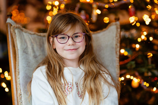 Smiling child, happy little girl relaxing in a chair beside a Christmas tree with warm lights, capturing a cozy real life family moment