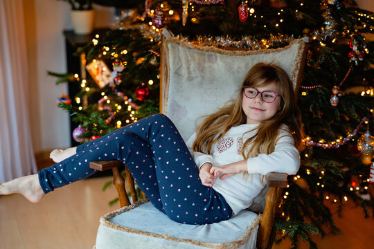 Smiling child, happy little girl relaxing in a chair beside a Christmas tree with warm lights, capturing a cozy real life family moment