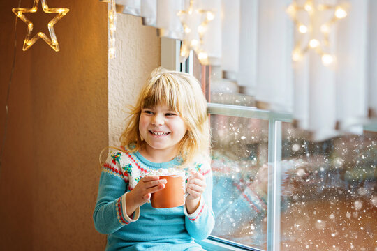 Little preschool girl holding cup with hot chocolate with marhsmallows. Happy child drinking sweet cocoa by window with Christmas lights in winter. Cozy family celebration of xmas.