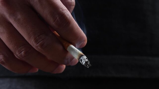 close up man holding smoking a cigarette in hand. smoke spread. dark background.