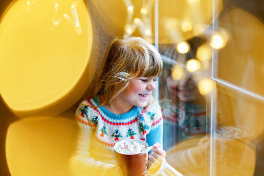 Little preschool girl holding cup with hot chocolate with marhsmallows. Happy child drinking sweet cocoa by window with Christmas lights in winter. Cozy family celebration of xmas.