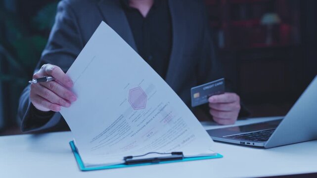 Businessman holding credit card reviews financial document beside laptop computer, managing online payment transactions inside dark office setting late night.