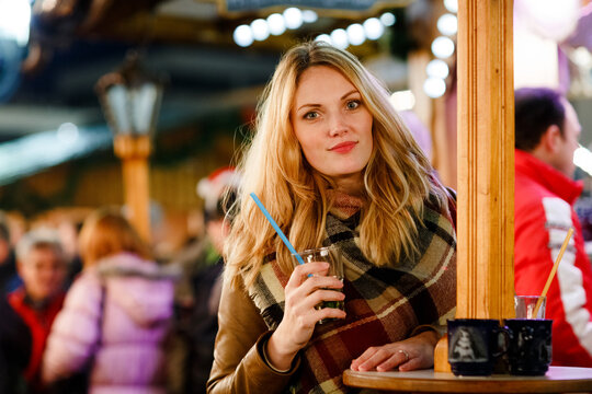 Happy young woman drinking hot punch, mulled wine on German Christmas market. Happy person in winter clothes with lights and friends on background. Family, tradition, holiday concept.
