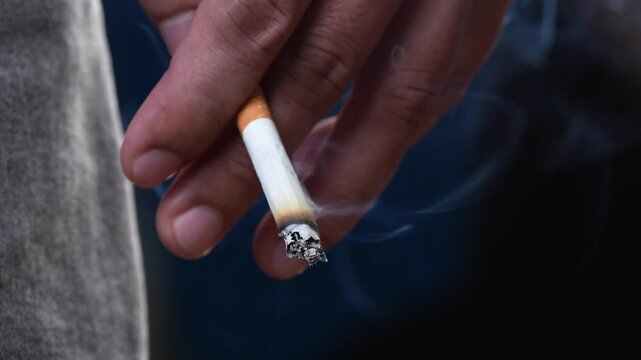 close up man holding smoking a cigarette in hand. smoke spread. dark background.
