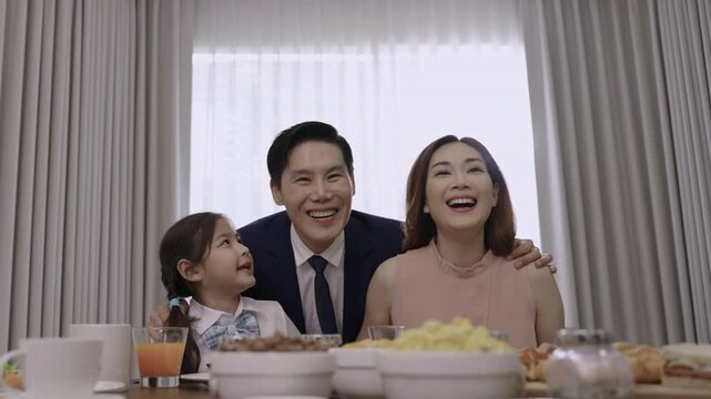 Happy Asian family relationships at the dining table in the morning, with father in a suit, mother, and a child girl smiling, teasing, and bonding before eating breakfast together in kitchen at home. 