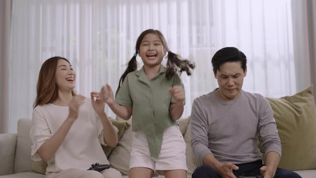 Happy family day concept. Asian father, mother, and daughter sitting using joystick and playing video game together having fun and cheerful on sofa in living room at home while child girl she winner. 