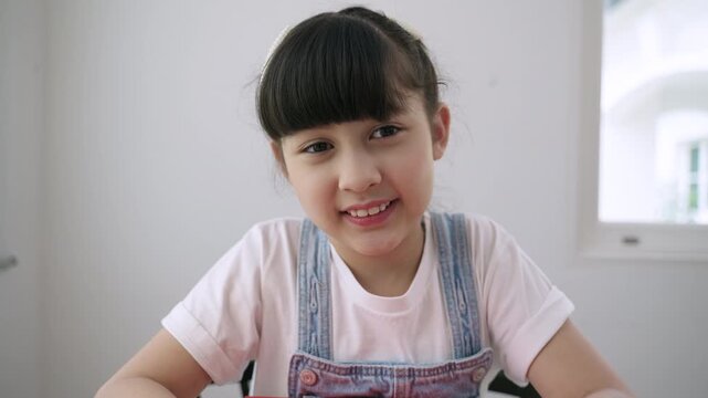 Cute Asian girl student sitting at desk is looking at the webcam while talking to computer camera and video calling with a remote teacher for online learning at home. Homeschooling education concept. 