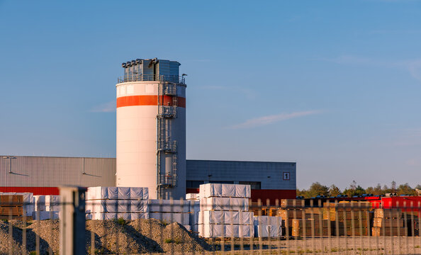Industrial silo and lumber yard with stacked wrapped timber and warehouse buildings behind a wire fence in warm evening light against blue sky, wood industry, copy space