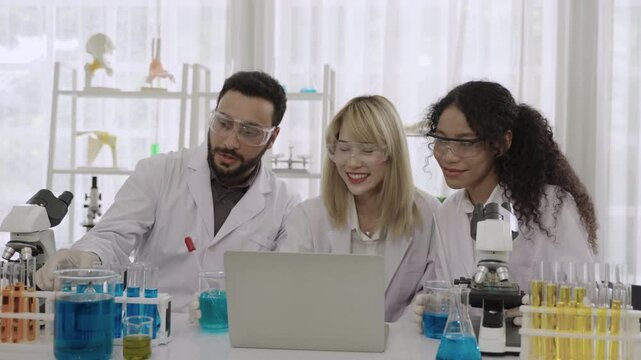 Group of happy men and women ethnic diversity, scientists and researchers wearing lab coats and goggles sitting and using a computer laptop for an online conference meeting together in the laboratory.