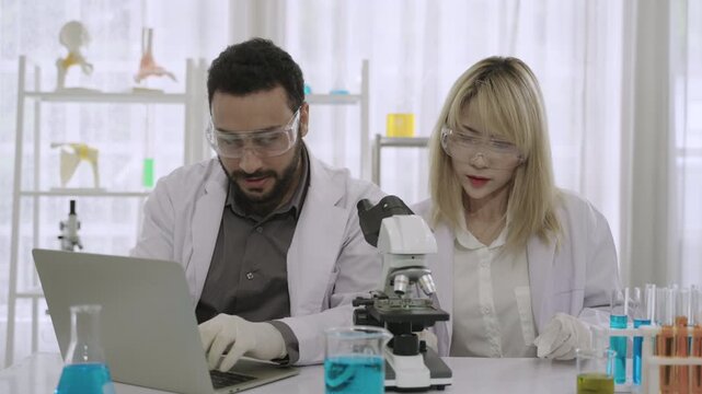 Team happy professional scientists and researchers in lab coats using a microscope and a laptop while conducting chemistry experiments with liquid in test tubes and beakers in the science laboratory. 
