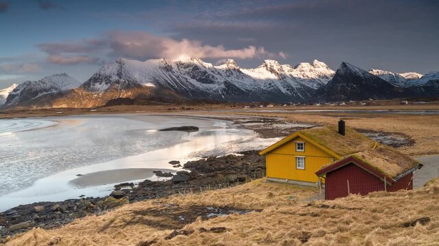 View of traditional yellow and red wooden houses with grass roofs situated by the coast with snow-capped mountains in the background in Reine, Nordland, Norway.