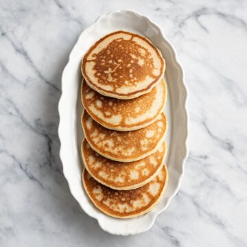 A stack of golden brown homemade pancakes served on an elegant white scalloped platter on a marble surface