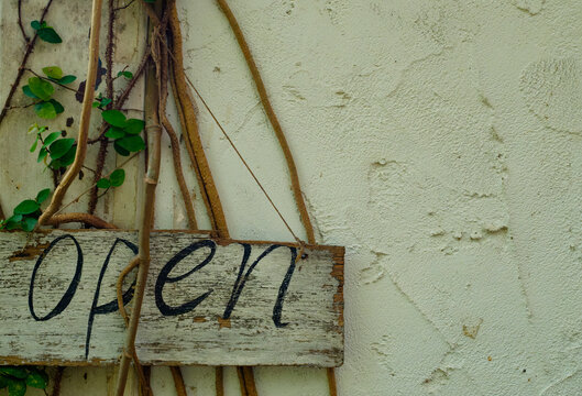 Rustic wooden open sign hanging on a weathered white stucco wall with climbing green vines