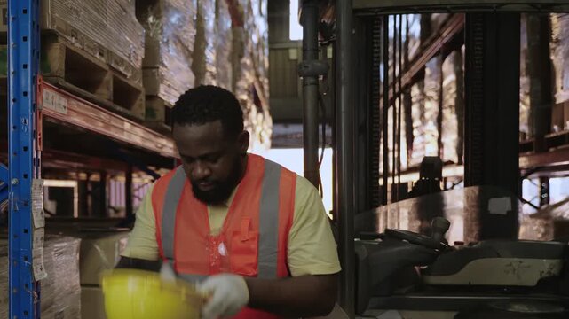 Professional African American male forklift driver wearing a yellow safety helmet, working by driving to move pallet cargo boxes on a shelf for delivery in a logistics distribution warehouse factory. 