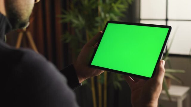 Digital nomad lifestyle and remote work concept, man holding tablet with green screen for mockup, handheld over the shoulder shot in home office