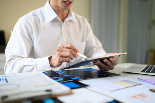 Businessman Reviewing Data Charts on Tablet with Stylus at Office Desk Surrounded by Printed Reports
