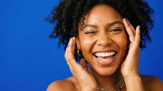 Joyful Black woman laughing and winking in an extreme close-up portrait against a vivid cobalt-blue background, energetic commercial beauty photography, hands framing her face, warm luminous skin,