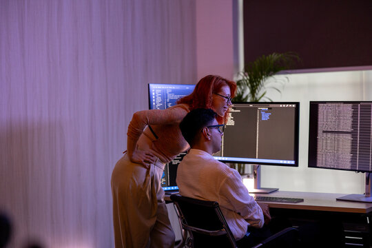 Two focused programmers collaborating on code on multiple computer monitors while working late in a dark office with neon lighting.