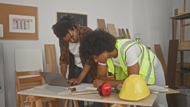 Two men carpenter partners lean over a laptop and blueprint while one writes notes with a pencil, high visibility vest and hardhat on table at workshop; focused teamwork.