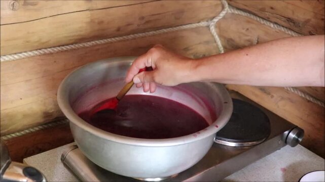 A woman makes redcurrant jelly, stirring the mixture with a spoon in a metal basin on an electric hot plate in a country house. Homemade berry preserves &ndash; horizontal video, close-up, woman's hands