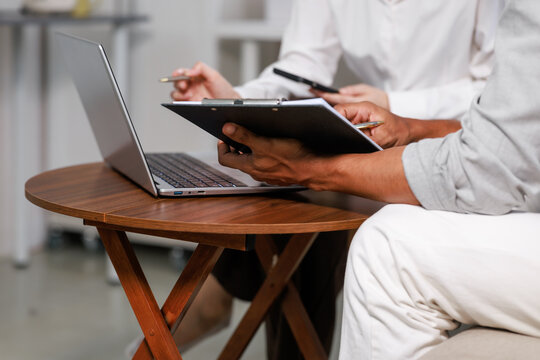 Close view of two coworkers reviewing paperwork on a clipboard beside an open laptop at a small wooden table in a modern office.